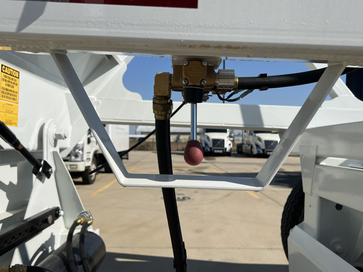 Trailer frame with hydraulic hose and valve assembly, gold fittings, black hose, pink protective cap; surrounding white metal structure and tire visible.