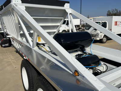 Dump trailer showing tri-axle air ride suspension with skeleton frame and large hopper in front, white-painted metal with black air tank and blue hoses.