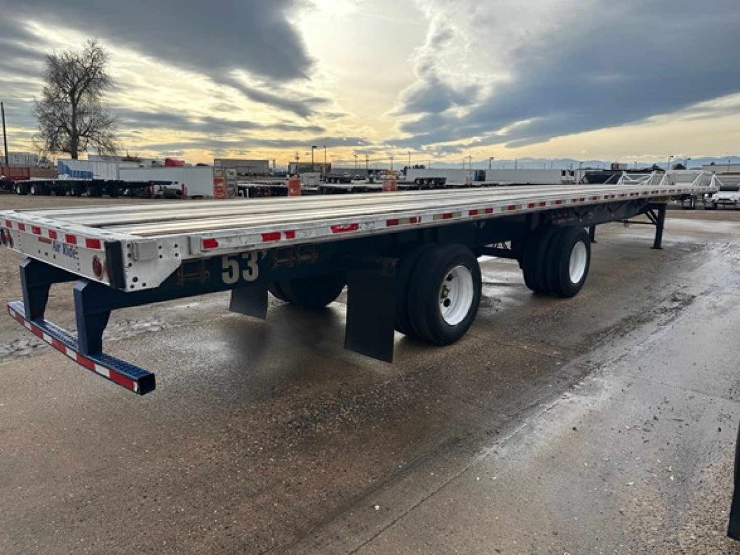 Flatbed trailer in a lot, viewed from rear-left side. White aluminum frame with reflective red/white stripes along sides. Dual rear axles with black tires. Some text and numbers painted on frame and mud flaps.