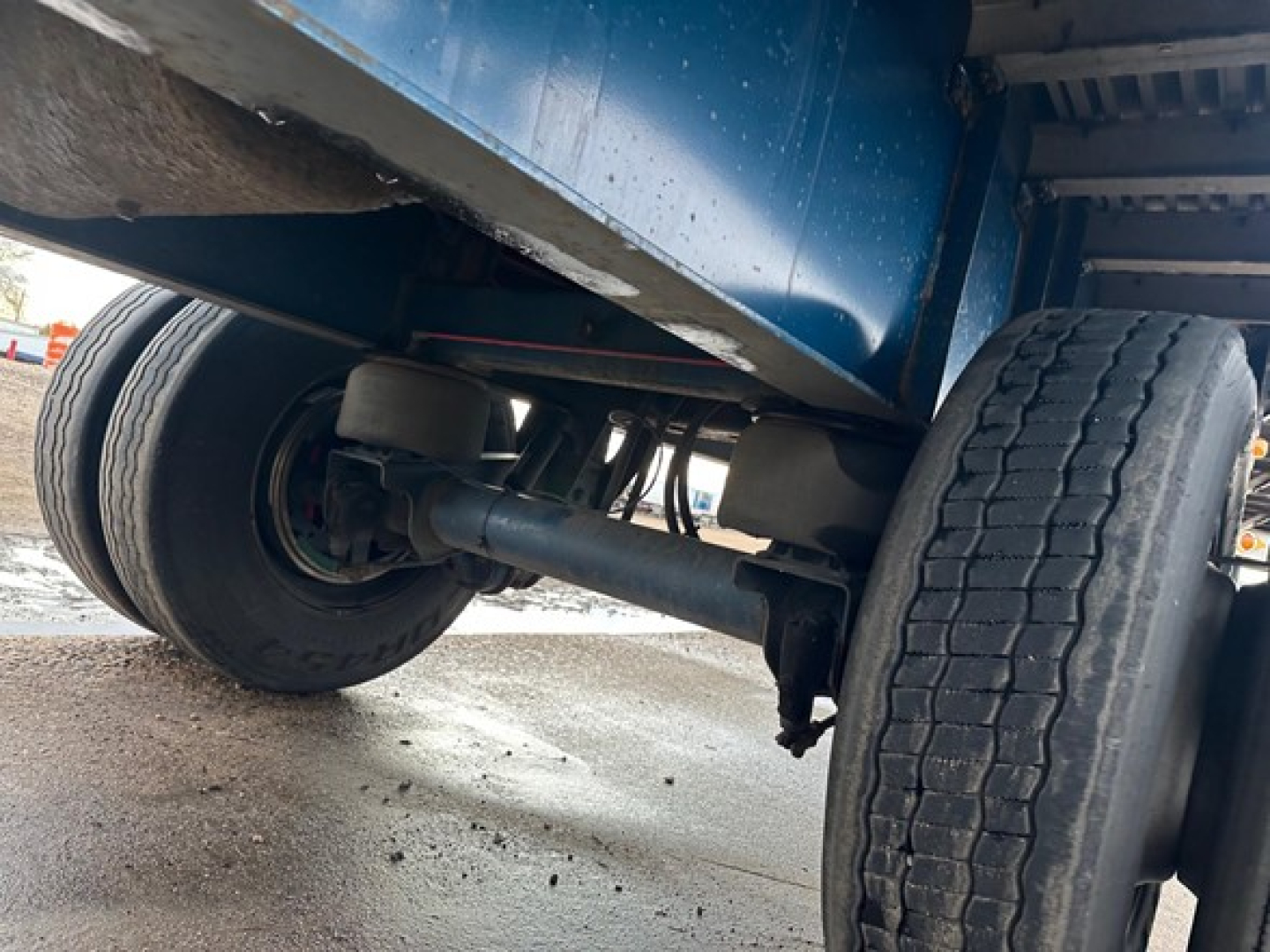 Undertruck view of a flatbed trailer showing dual rear axles and tires, with undercarriage components and suspension visible.