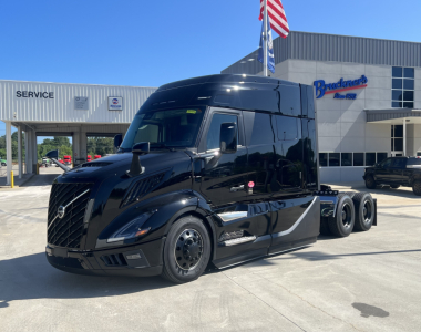 Volvo VNL64T840 truck in black with sleeper cab, front view, three rear axles, dealership building in background.