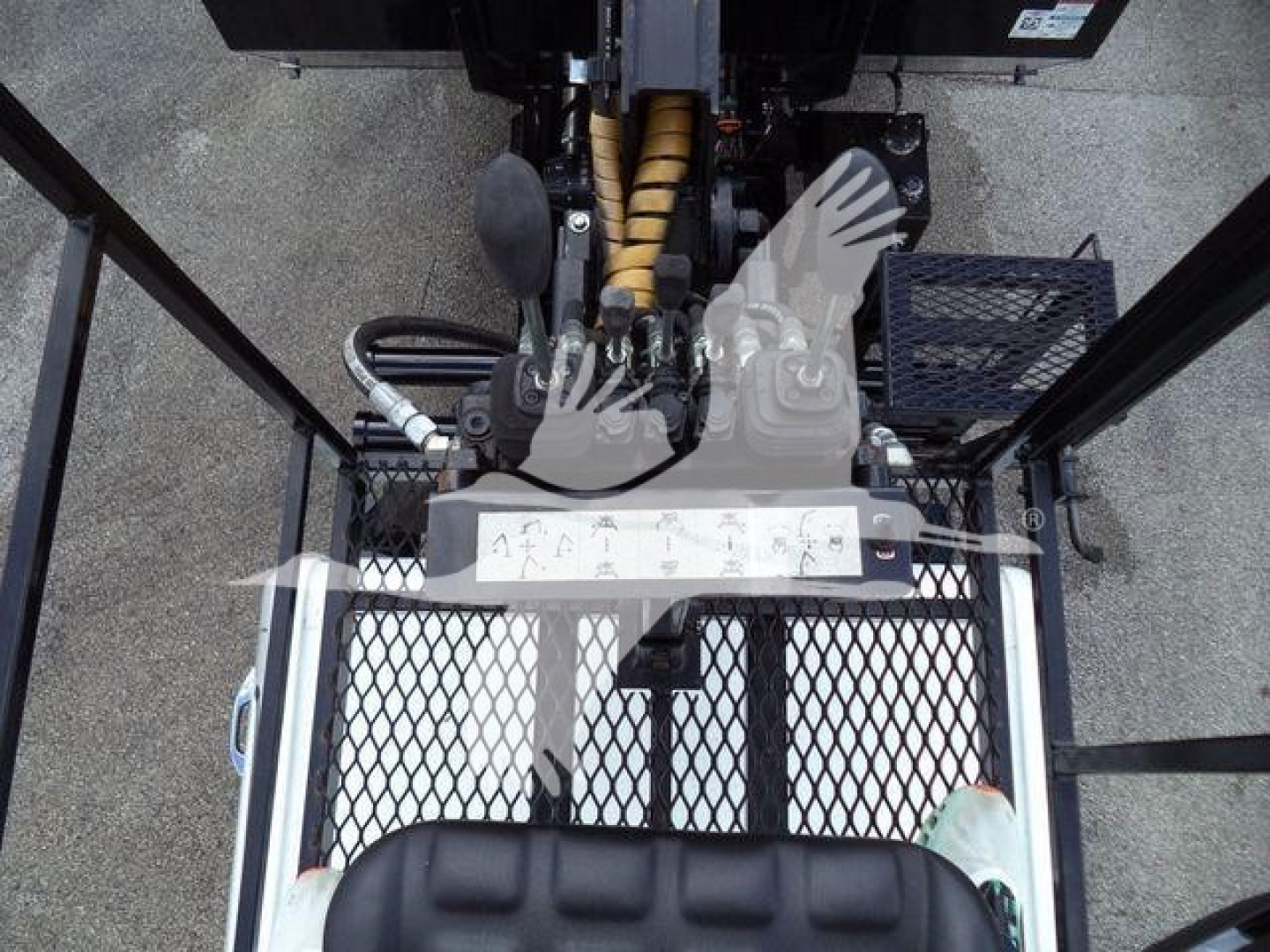 Truck cab view showing equipment under front loader: hydraulic controls, metal mesh platform, pedals, and a labeled control panel.