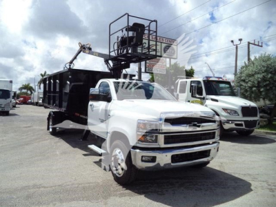 Truck bed with a raised lift, white Chevrolet Silverado 6500HD in foreground, black utility body and crane behind, dealership lot