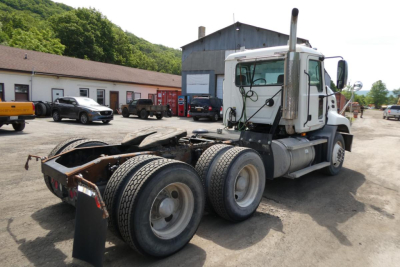 Truck chassis and cab of a 2007 Mack CXN613 daycab with exposed frame and dual rear axles, white cab, large vertical exhaust stack, service area in background.