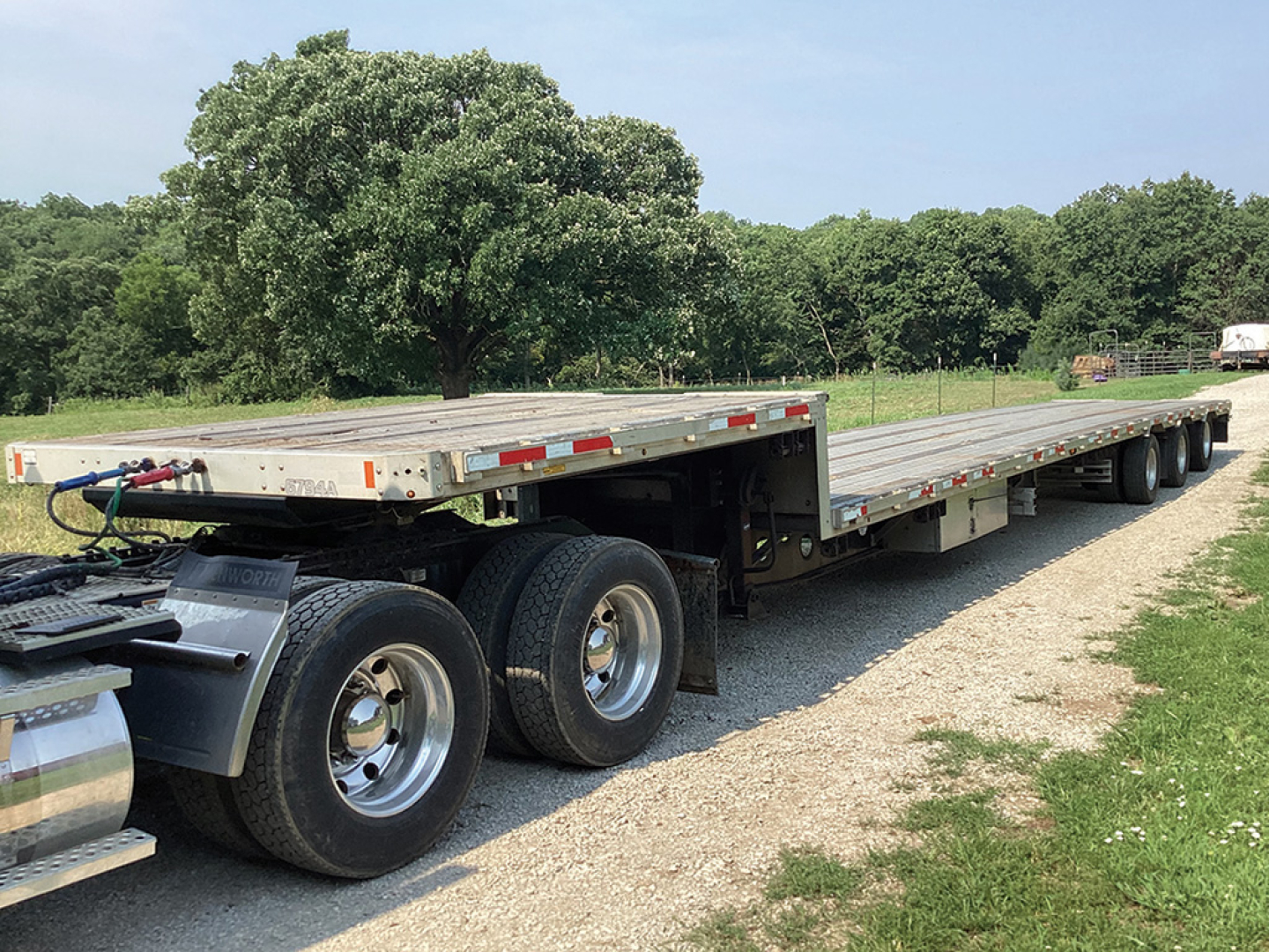 Long drop deck trailer with multiple axles in a field. Visible text includes small labels on side and near the hitch, front corner features chrome fuel tank.