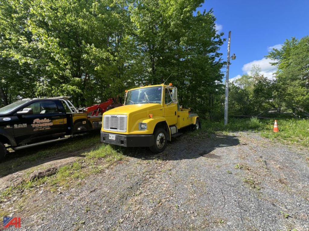 Front view of a yellow 1998 Freightliner FL50 wrecker truck parked on a gravel lot with trees in the background.