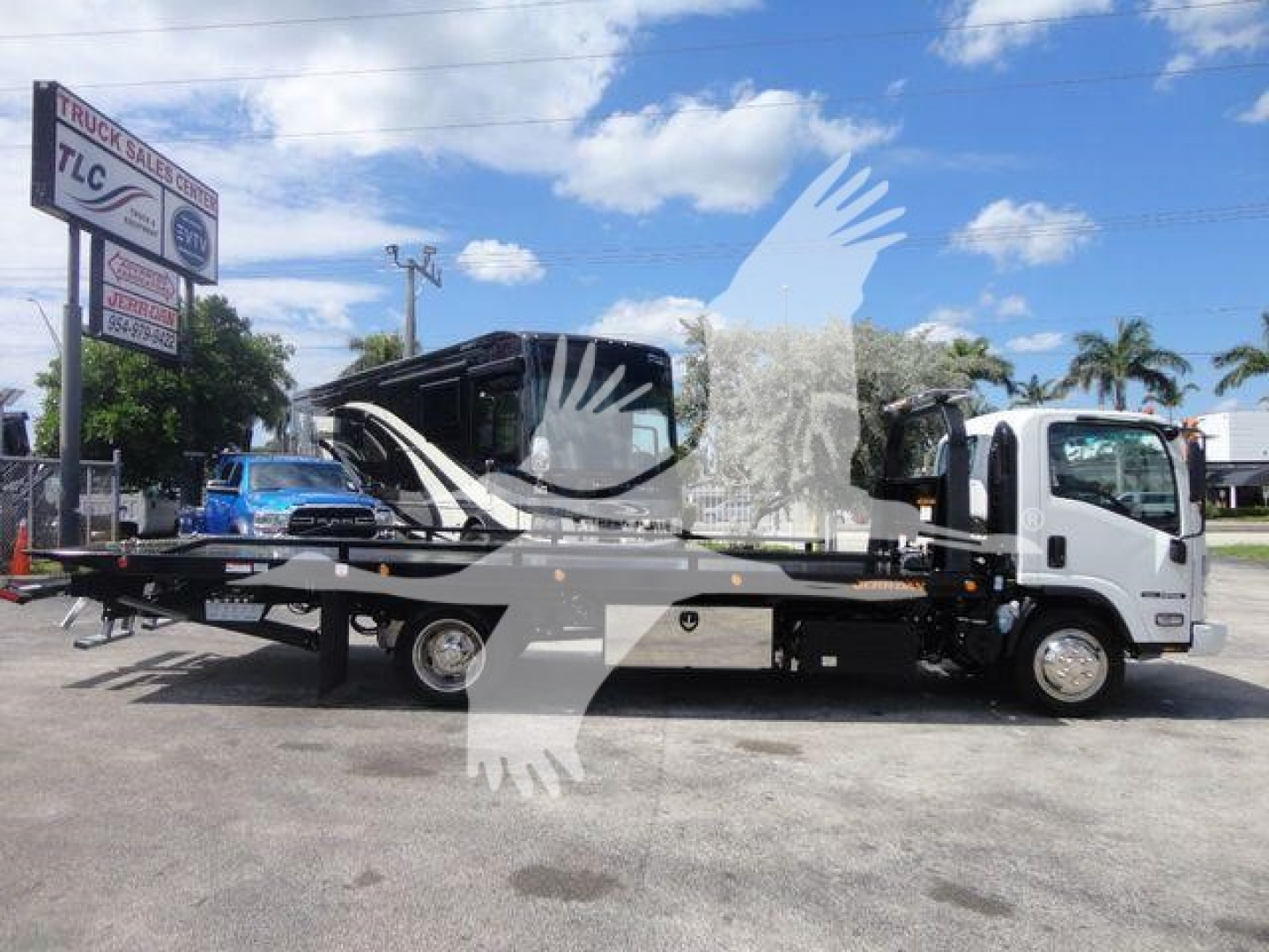 Isuzu NPR rollback tow truck side view showing flatbed with hydraulic ramps and cab in white, dealership signage in background.