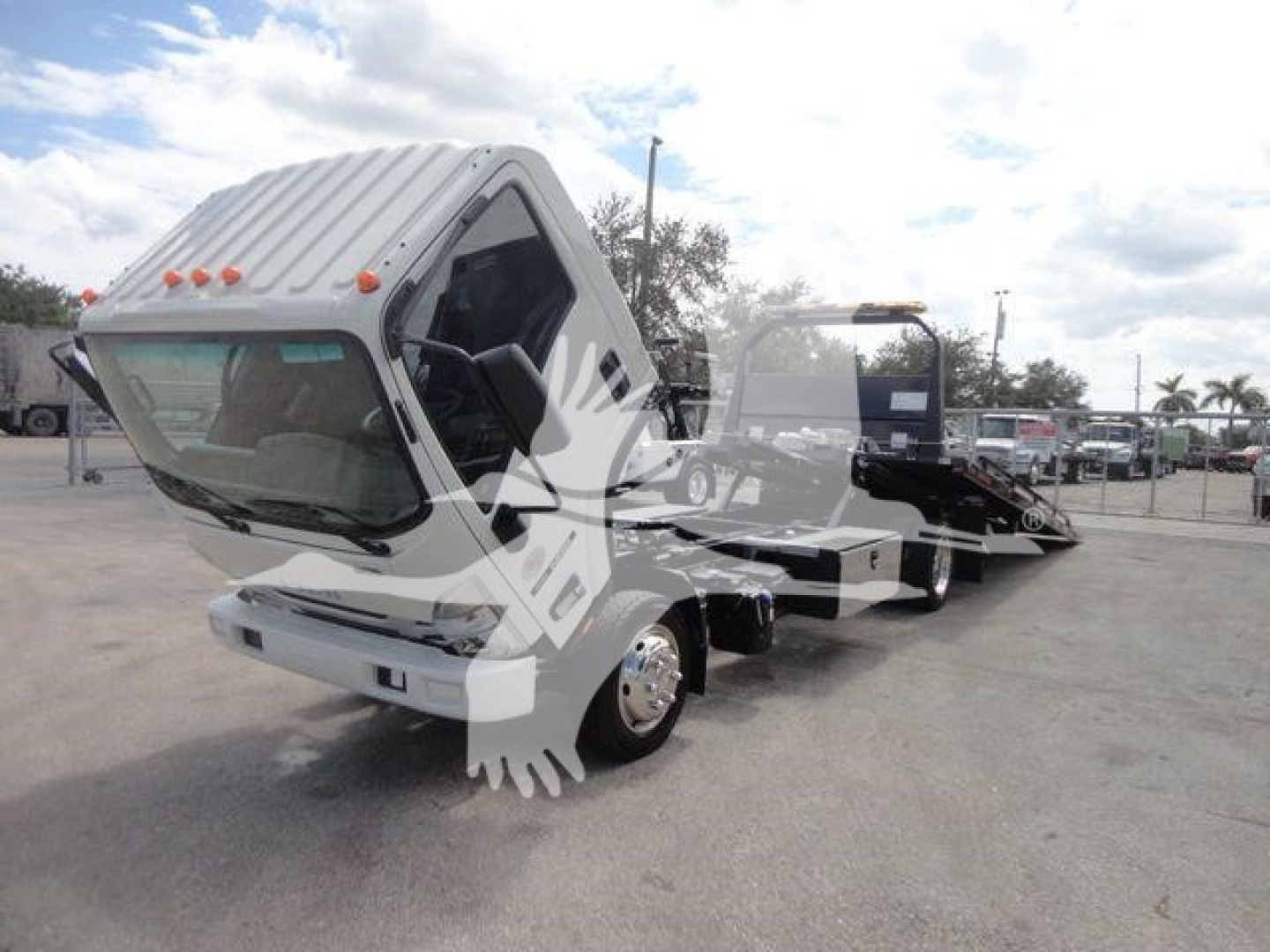 Isuzu NPR/NRR cab-front and chassis with rollback truck body, white cab, tilted forward for cooling. Dashboard and windshield visible, multiple trucks in lot.