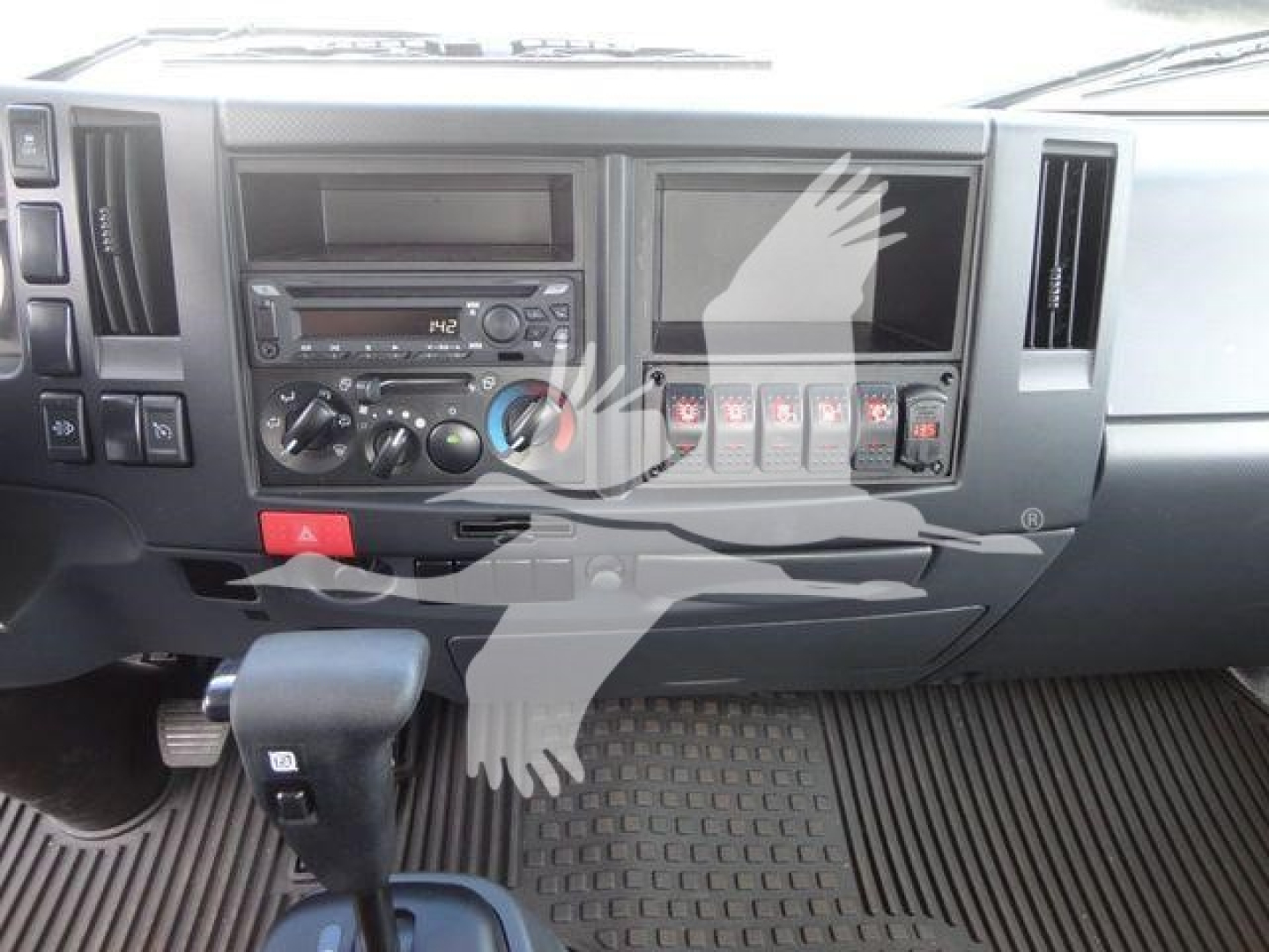 Truck dashboard and gear selector of a 2023 Isuzu NPR/NRR style cab showing center console, radio, climate controls, and vertical switch bank.