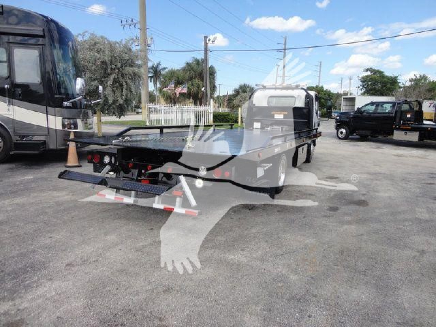Flatbed rollback truck in a parking lot, Isuzu NRR chassis with open flatbed and rear ramp; other trucks visible in background.