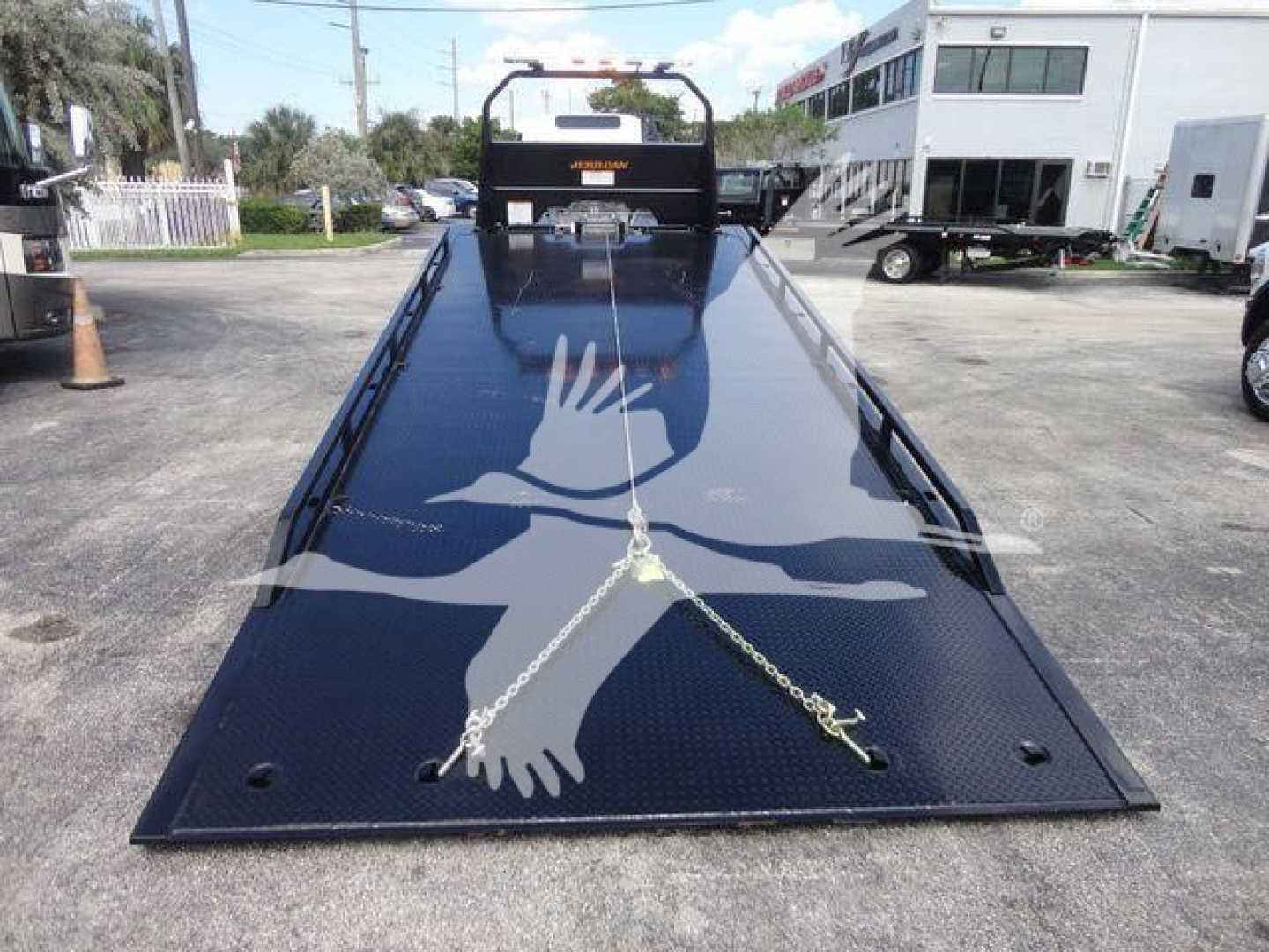Flatbed tow truck bed showing black textured steel surface with chains and ramp, Isuzu logo on headboard in background.
