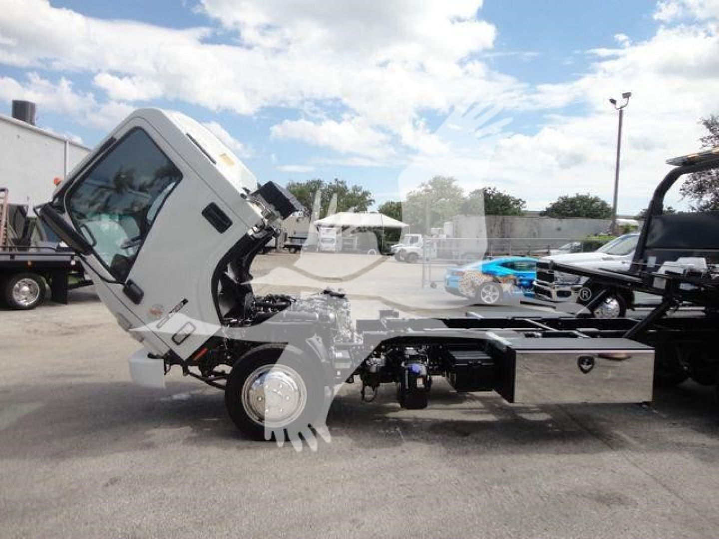 Isuzu NRR rollback truck chassis with cab tilted forward, side view showing engine area and fuel tanks, parked lot with other trucks.