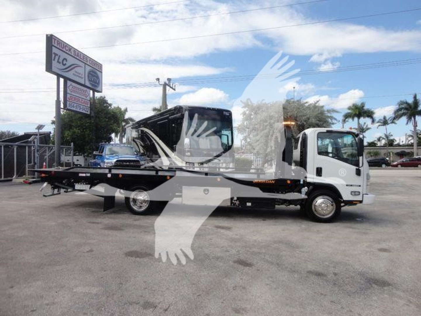 Isuzu NPR/NRR tow truck with a flatbed, side view showing the cab and lifted bed; dealership sign in background reads TLC Truck Center.