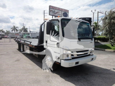 Isuzu NPR/NRR flatbed tow truck in a dealership lot viewed from front-left, displaying rollback bed and cab; signage in background.