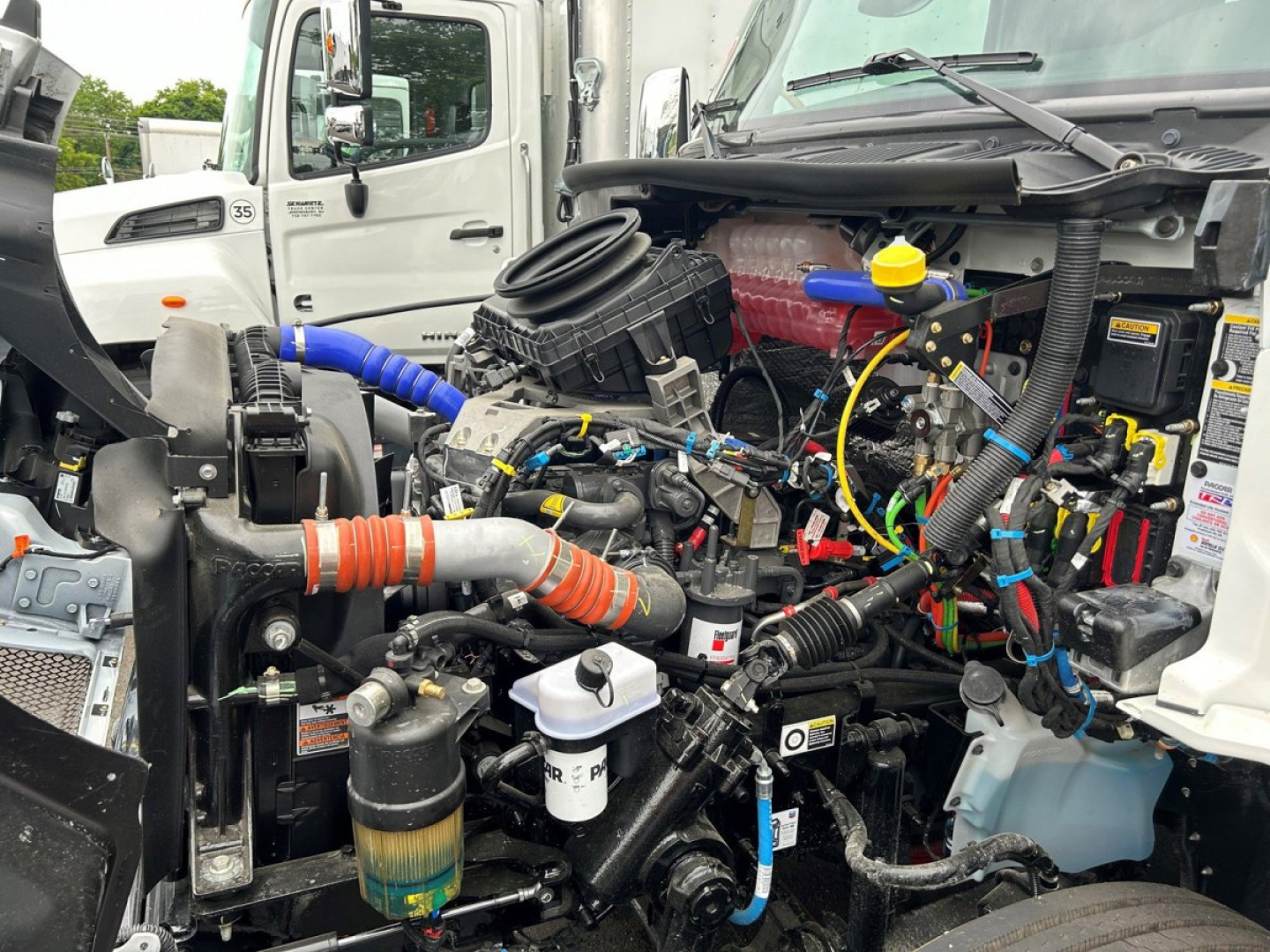 Truck engine bay with exposed 6-cylinder diesel and wiring harnesses; visible coolant reservoir, colored hoses (orange, blue), yellow dipstick, and various sensors; front of Kenworth T280 tow truck drivetrain.
