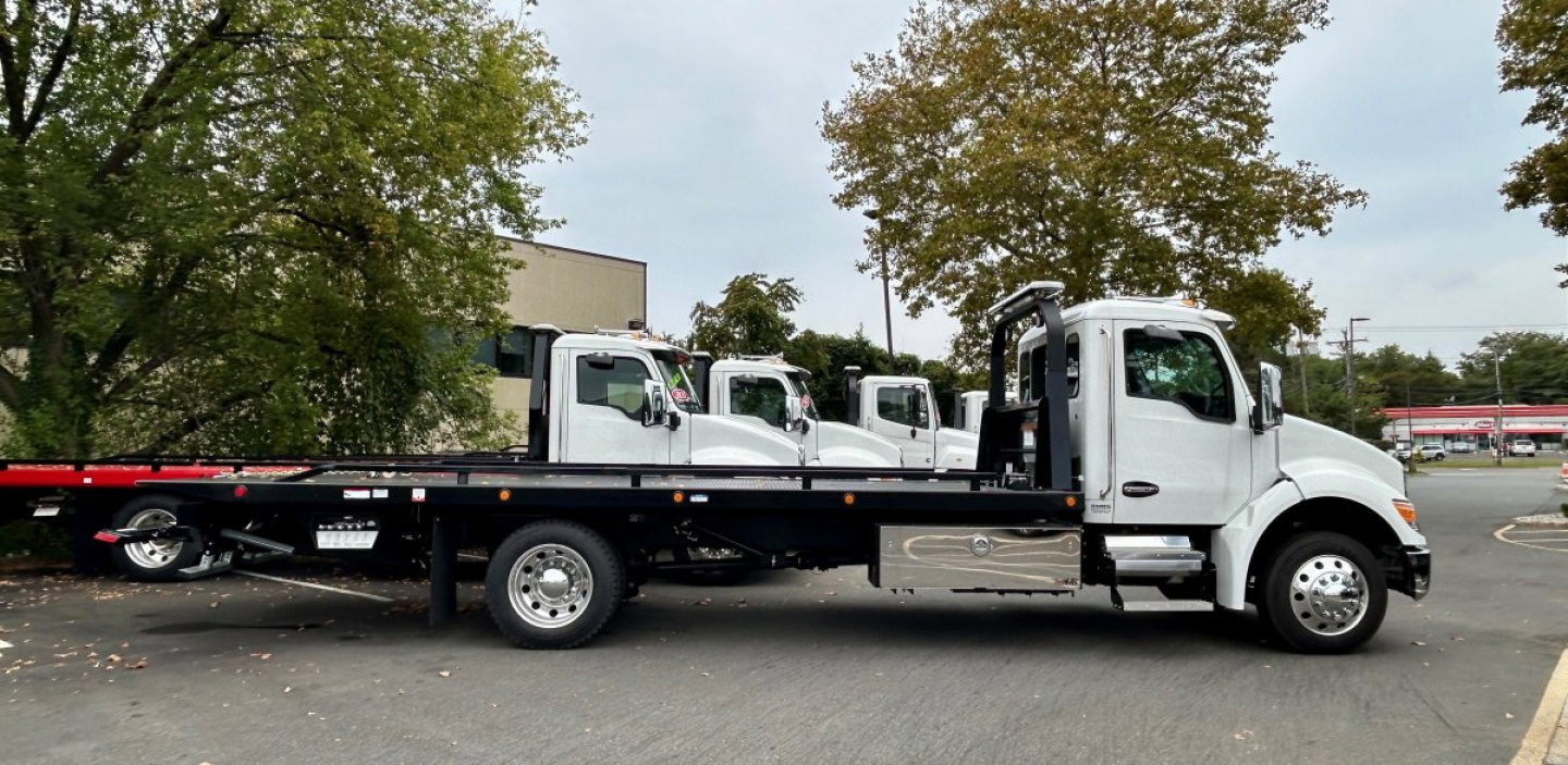 Tow truck flatbed showing Kenworth T280 cab and flatbed with multiple auxiliary tow trucks in background.