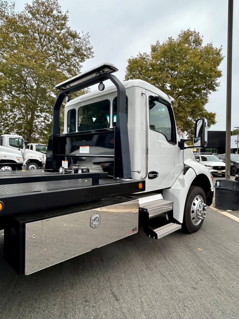 Kenworth tow truck cab with flatbed, side steps, and rear recovery equipment visible in a parking lot.