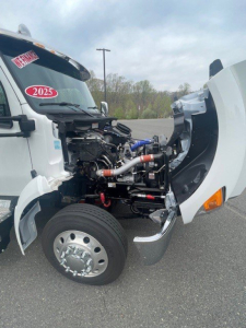Kenworth T280 front engine bay with exposed diesel engine and intake components, white cab, 2025 badge on windshield, open hood showing hoses and metal piping.