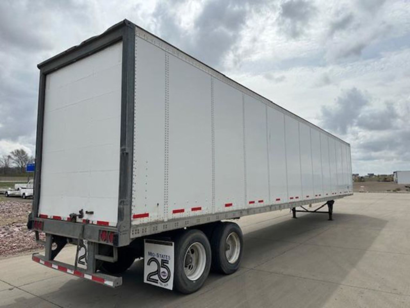 Trailer side view showing a long white box trailer with dual rear axles and rear doors, under bright cloudy sky.