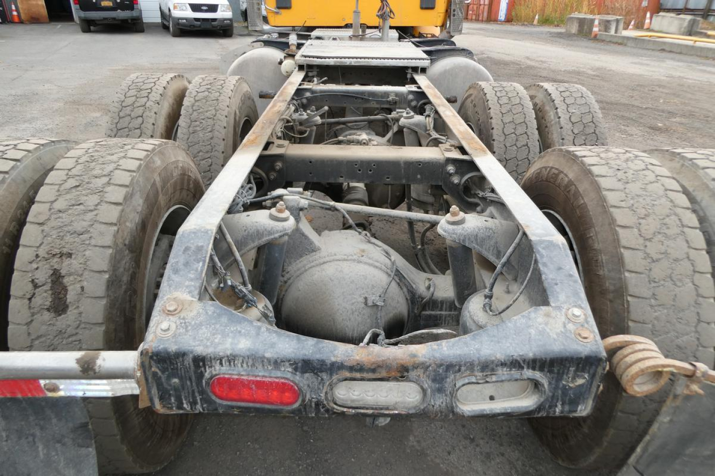 Rear view of a Peterbilt truck chassis showing dual rear axles, leaf springs, differential, and frame with faded mud; red and white rear lights and a hitch area visible.