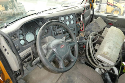 Peterbilt truck cab interior showing steering wheel, gauge cluster, and exposed wiring with a large battery or control module strapped to the passenger seat area.