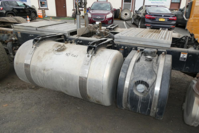 Truck chassis with fuel tanks and twin cylindrical tanks; visible labels include 'NO FUEL' on left tank. Background shows dealership lot with cars.