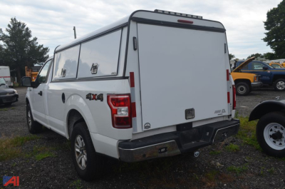 Ford F-150 pickup truck with a white utility cap/camper shell in rear, 4x4 badge visible, tailgate closed, rear bumper and hitch, outdoor lot.