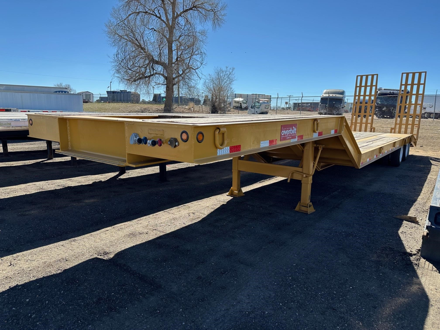Lowboy trailer with yellow deck showing control panels and dual ramps at rear. Visible labels include 'Great Dane' style sticker and side marker lights.
