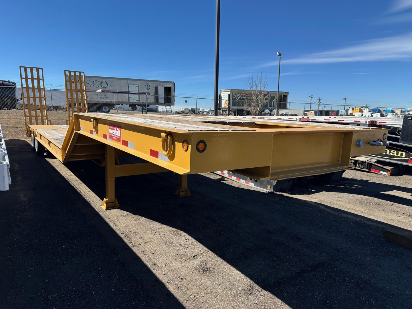 Lowboy trailer in a yard showing a long flatbed with yellow paint, ramps at rear, and safety decals on the side. Visible labels/markings include warning stickers and reflective tape, no legible serials.