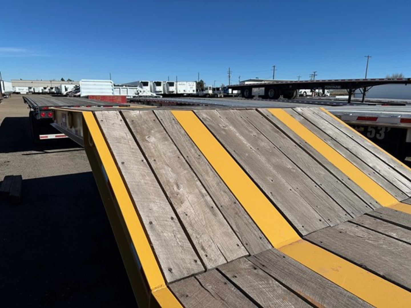 Equipment trailer on site with wood deck and yellow safety stripes, viewed from angle showing ramp and flatbed area.
