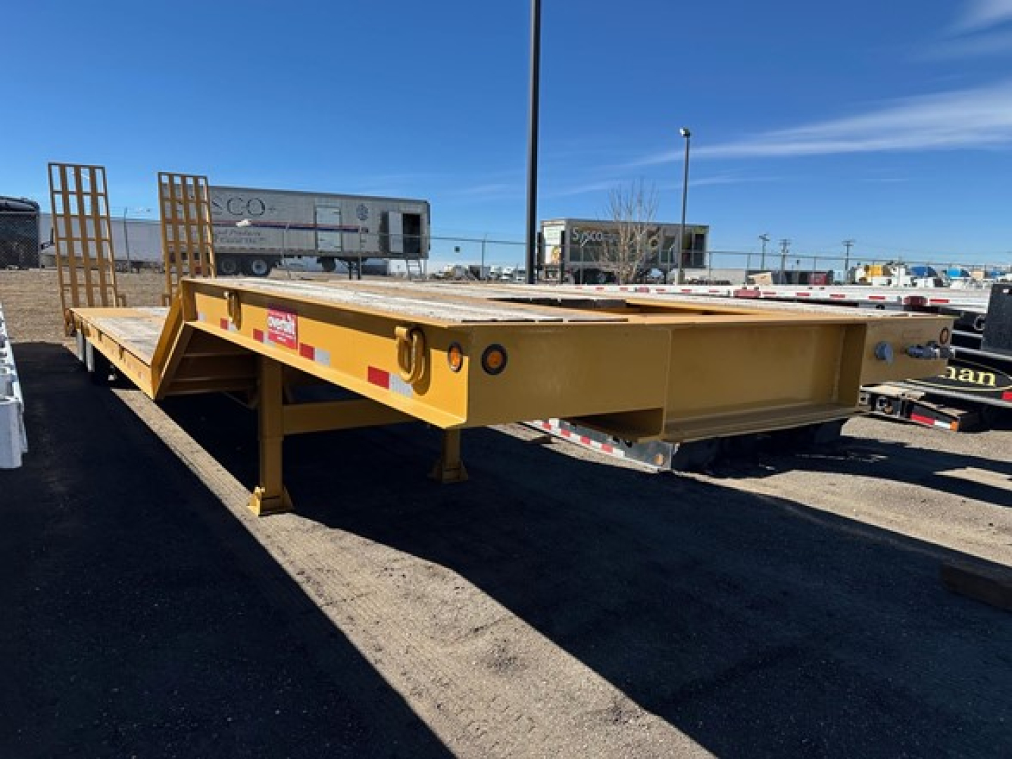 Equipment trailer with lowboy style, yellow-painted, flatbed with front ramps and side markers, showing sturdy frame and landing gear.
