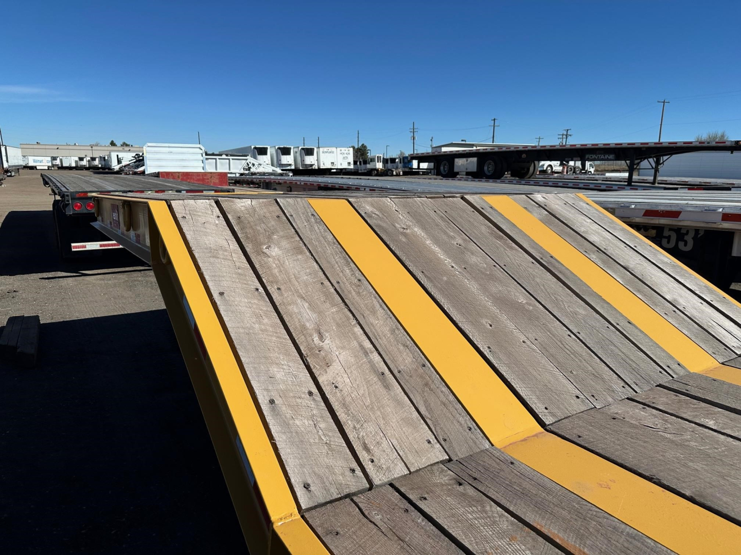 Lowboy trailer ramp with wooden planks and yellow safety stripes. Visible in the background are other trailers and a clear blue sky.