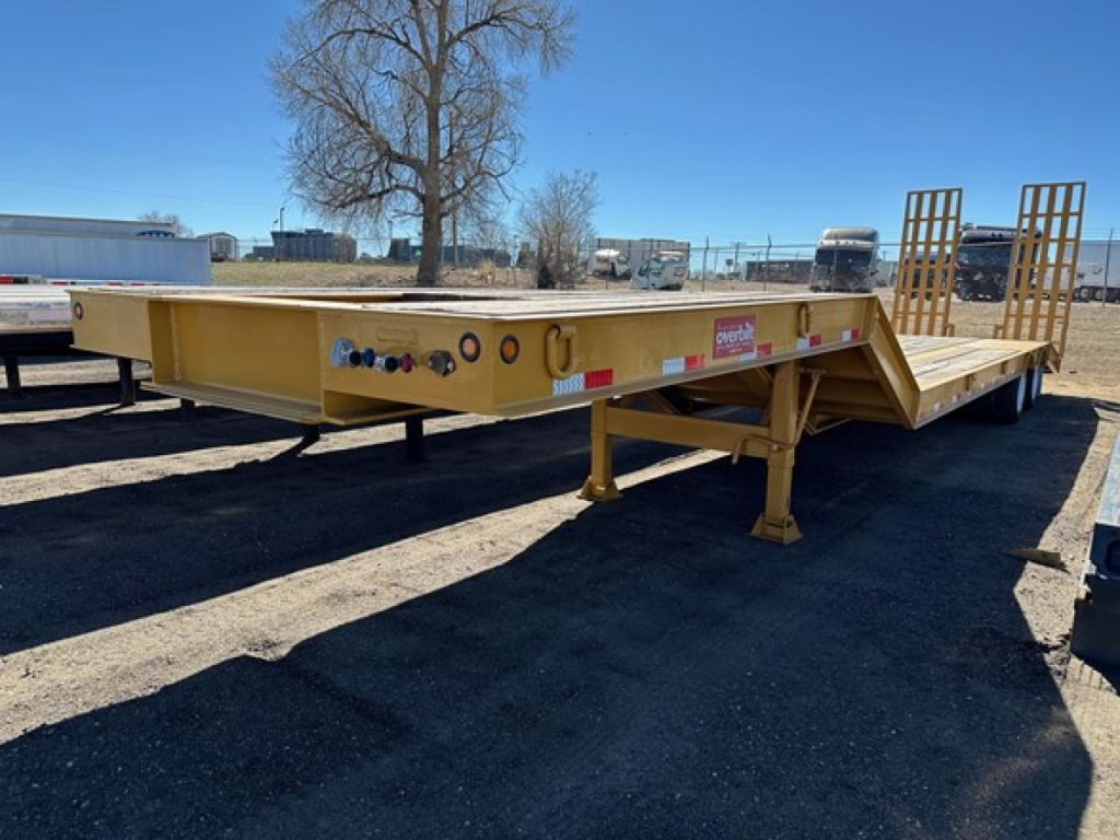 Equipment trailer showing lowboy deck, hydraulic air ramps, and loading ramps on rear; yellow painted trailer with wheel wells not visible.