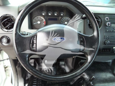 Ford steering wheel with dashboard gauges visible inside vehicle cabin. Center of steering wheel shows Ford logo; instrument cluster and controls visible.