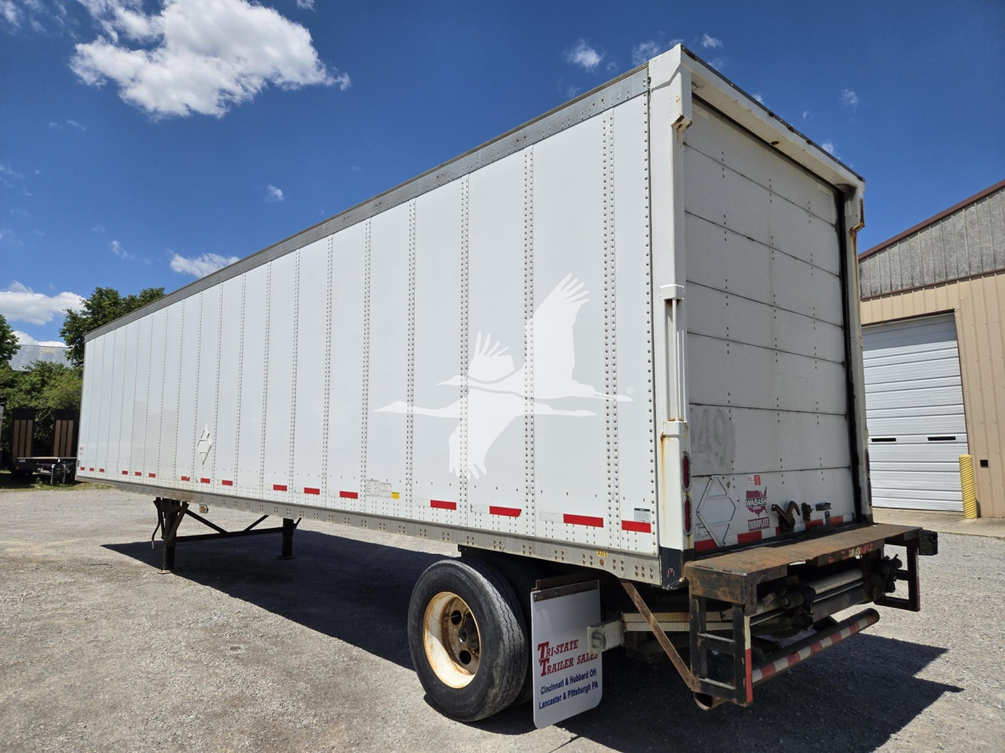 Trailer side view showing a white Wabash box trailer with bird graphic, rear doors, single axle, and jump steers in a yard.