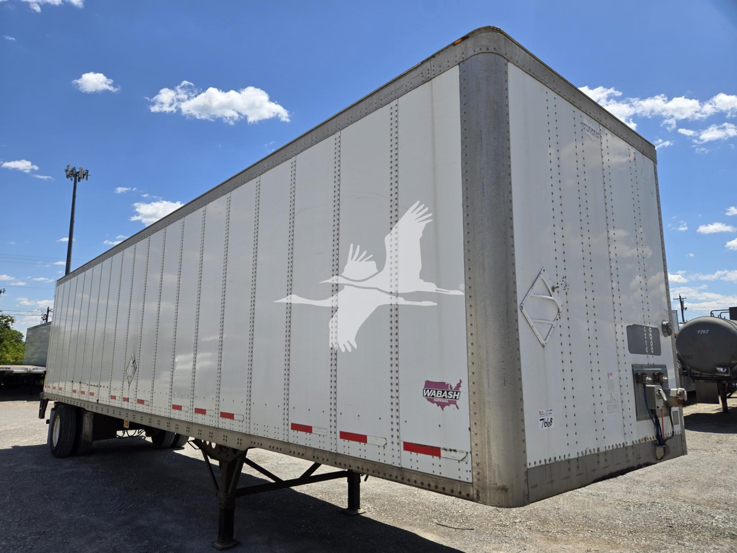 Trailer side view showing a white Wabash van trailer with a bird logo and reflective safety strips, quad rear doors, and landing gear visible under the empty trailer.