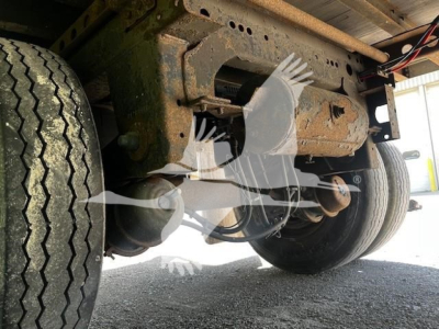 Underbody view of a flatbed trailer showing suspension and wheels. Visible text on the image includes no readable labels; the photo focuses on axles, leaf springs, and tires.