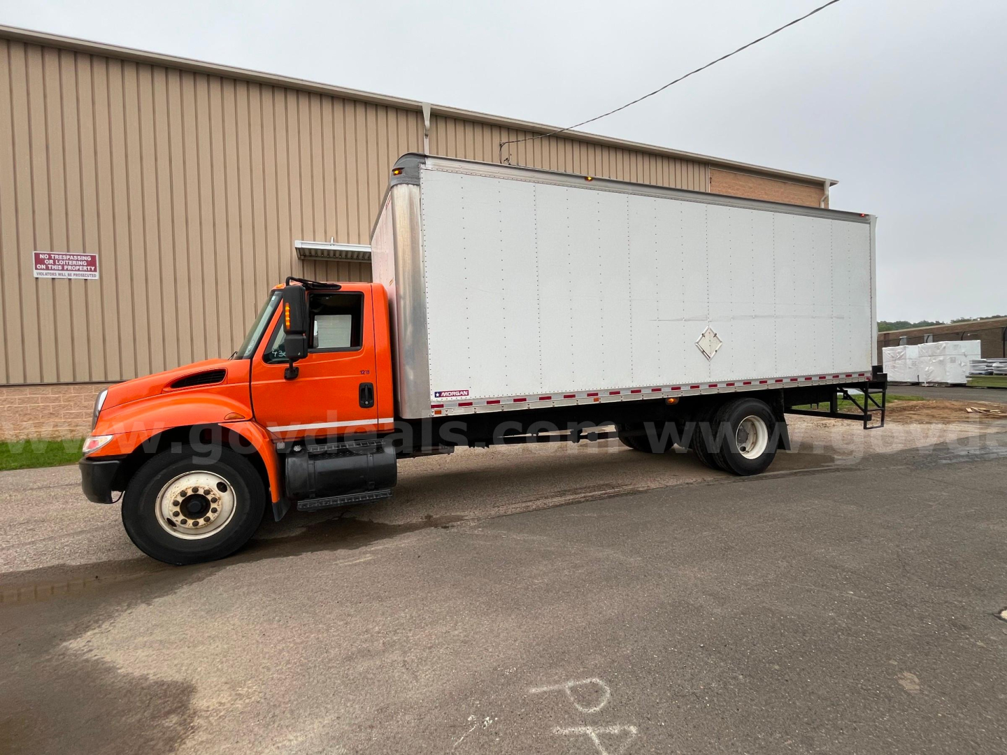 2004 International 4300SBA box truck in an outdoor lot, orange cab with a large white box cargo area. Side view shows dual rear wheels and a diamond-shaped hazard placard on the box.