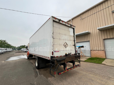 Box truck rear view showing white cargo box with diamond placard and rear bumper, loading dock building in background