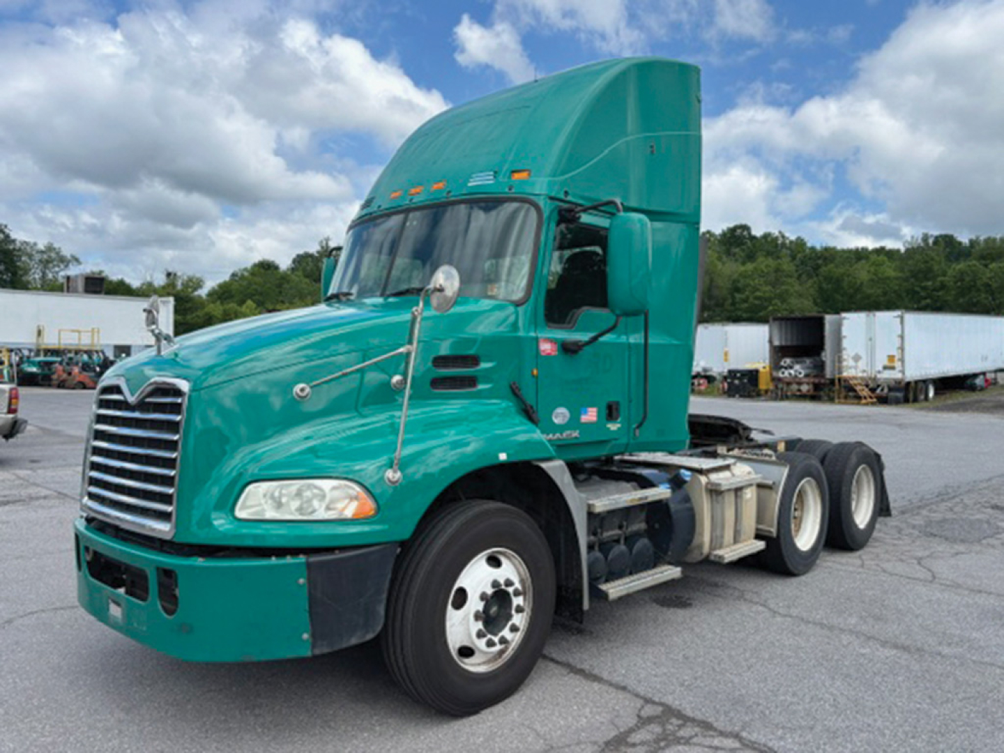 Truck cab in teal color with sleeper, badge on door, Mack CXU613 missing front bumper; left side shows fuel tanks and dual rear axles.