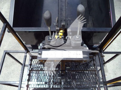 Controls and levers of a forklift-style control console with two joystick levers and a pedal area, seen from above, with some wired cables and a metal mesh platform.
