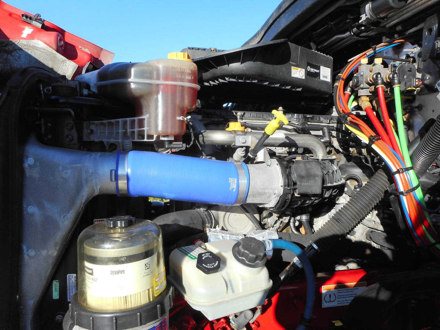 Engine bay of a Freightliner Cascadia showing blue intercooler hose and various fluid reservoirs and wiring Harness.