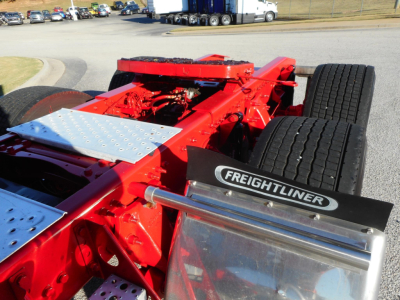 Freightliner chassis with red frame and dual rear wheels visible; metal steps and "FREIGHTLINER" badge on the side; part of a Cascadia sleeper truck under a bright outdoor lot.