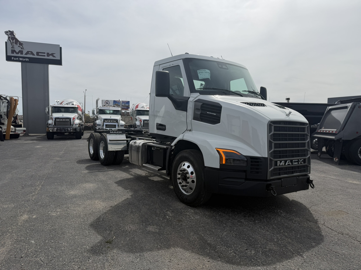 Mack AN64T truck cab in a dealership lot with white exterior, front grille, headlights, and Mack badge. Visible text includes MACK on the grille and dealership signage in the background.