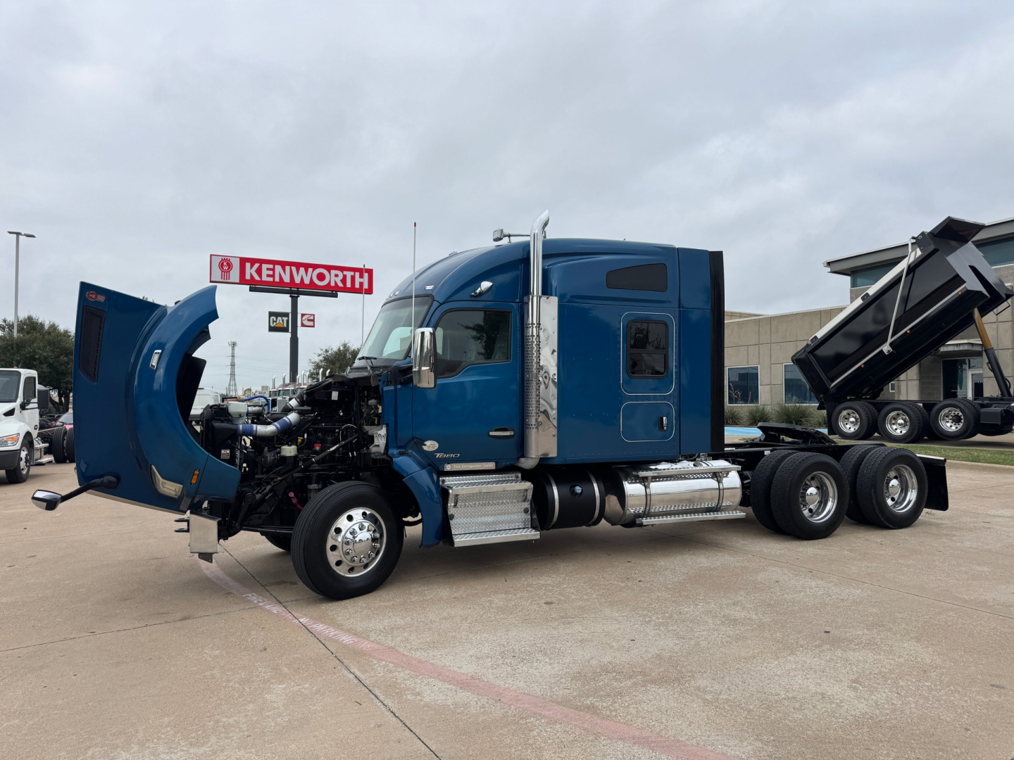 2019 Kenworth T880 with hood open showing engine bay side view. Blue sleeper cab tractor chassis with fuel tanks and chrome steps; Kenworth dealership sign in background.