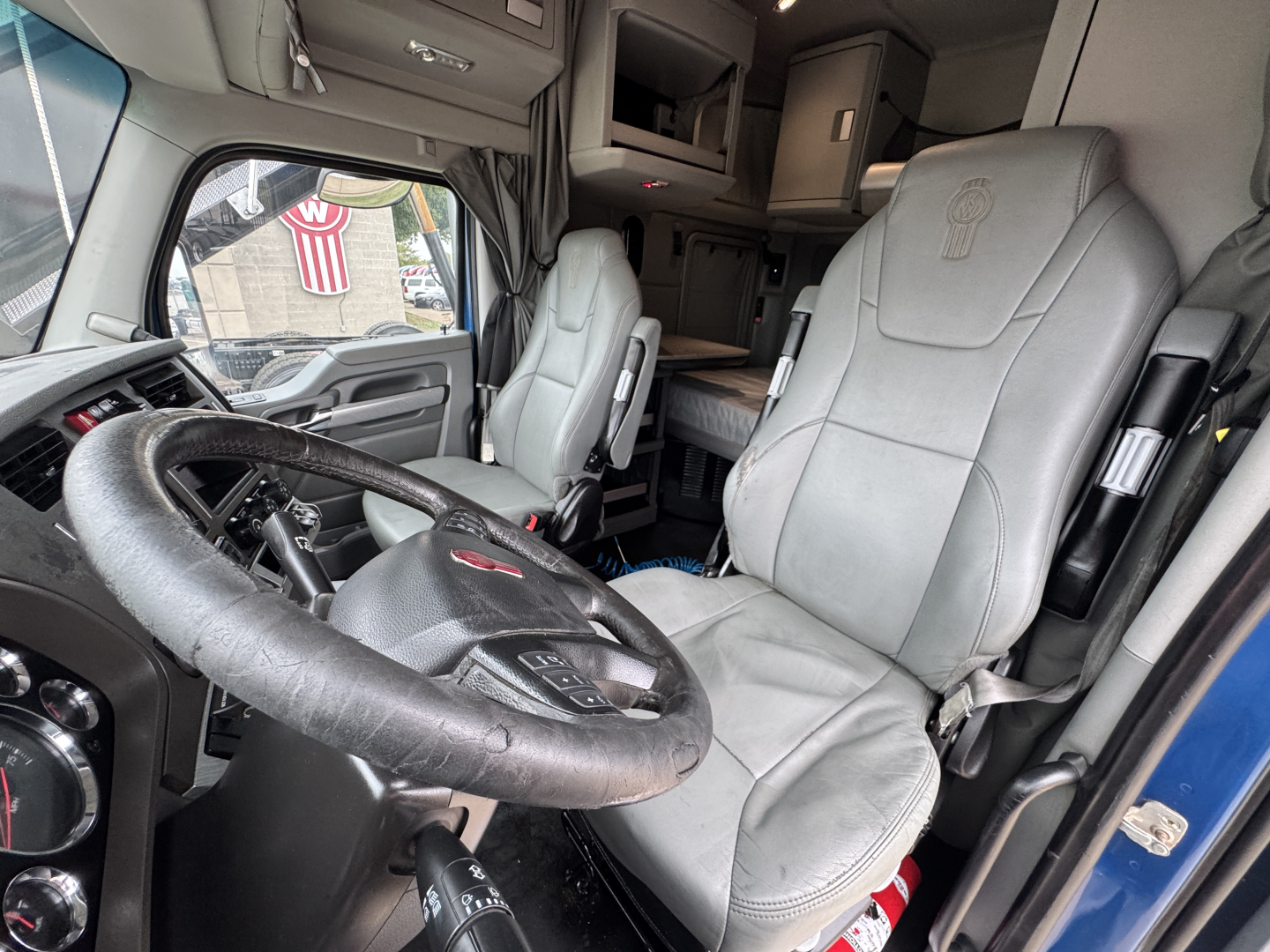 Truck cab interior with steering wheel, driver seat, and dashboard; Kenworth emblem on seat back; visible overhead sleeper area.