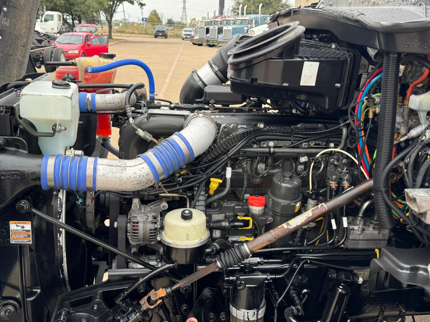 Kenworth T880 engine bay showing turbo intake and various hoses and wiring; blue silicone hoses, coolant reservoir, battery, and engine components visible.