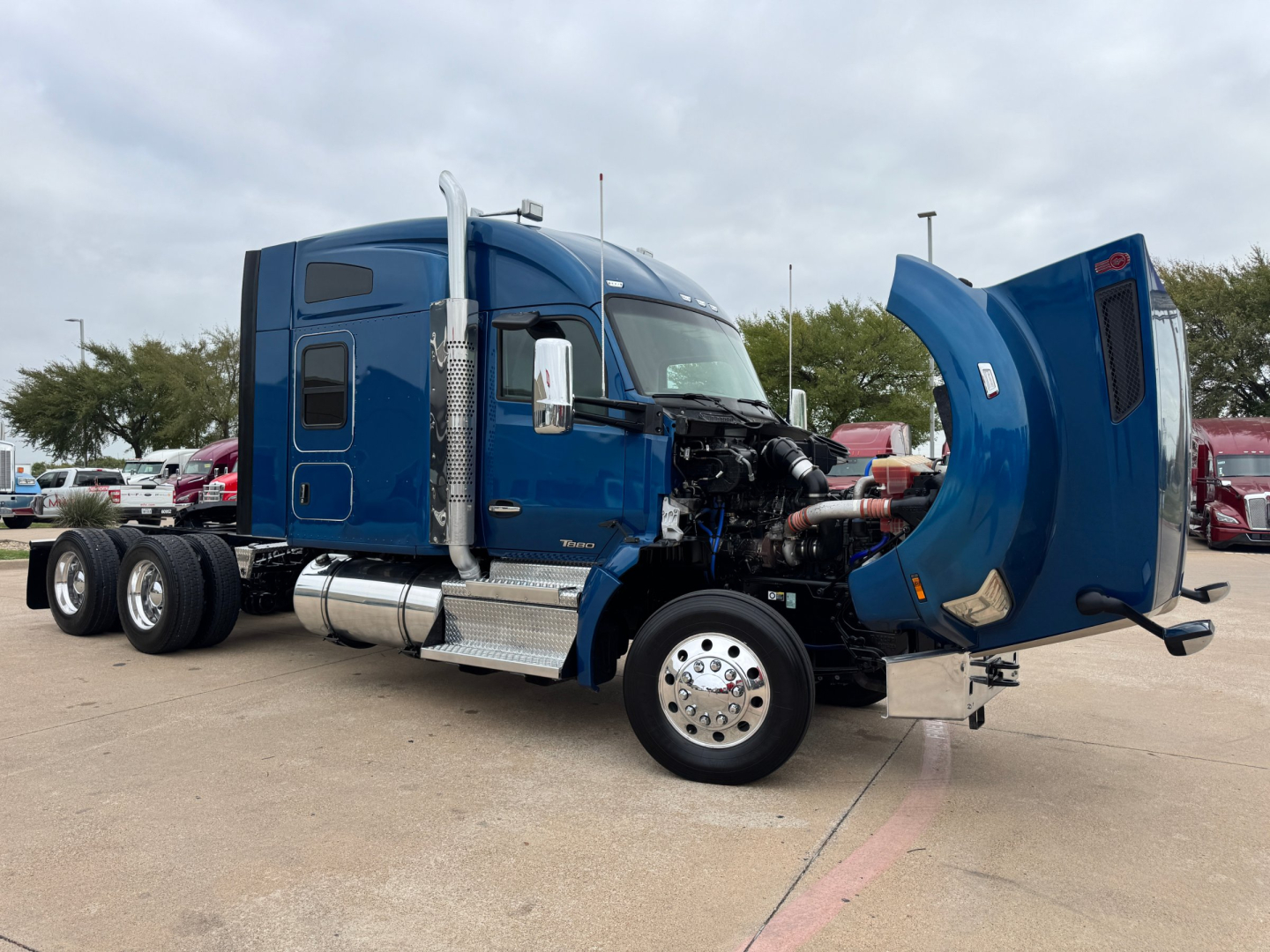 Kenworth T880 conventional sleeper truck with hood open showing engine bay and side exhaust, blue paint, chrome fuel tanks, dual rear axles.