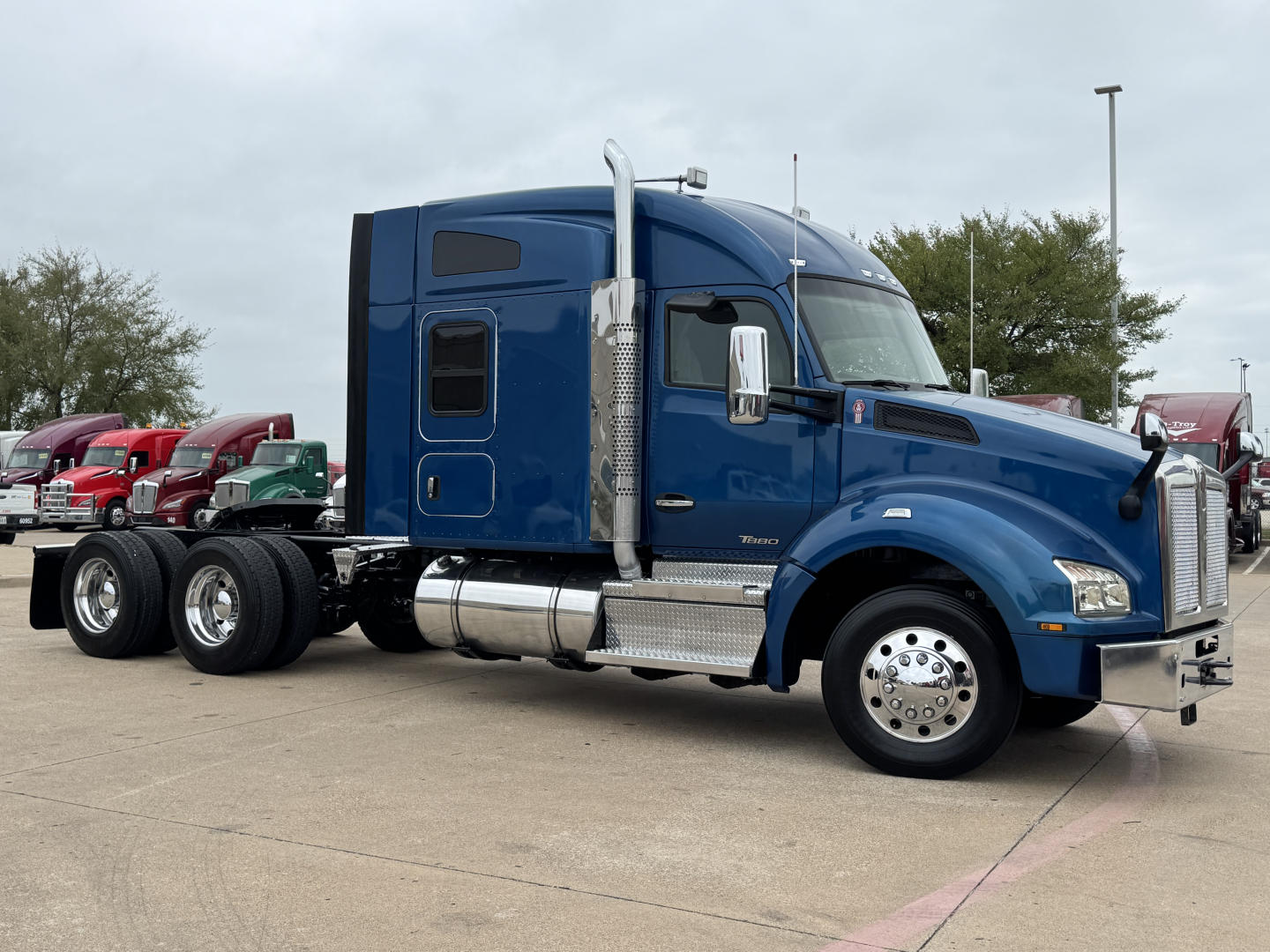 Kenworth T880 sleeper cab tractor in blue, side view showing chrome fuel tanks, steps, and dual rear axles with triple wheel configuration.