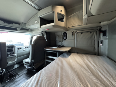 Kenworth truck cab interior showing sleeper berth, overhead storage, dashboard with steering wheel, and curtain. Visible text on equipment labels: none clearly legible; general interior components in gray.
